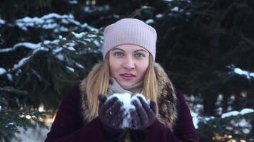 A beautiful happy girl in a light hat blows snow from her hands in the open air in winter having a good break up and smiling. video