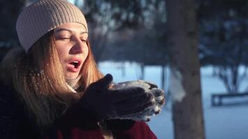 Close-up. A young cute girl in a light hat blows a pile with snow in the winter in the forest against the background of snow and sun having a good mood video