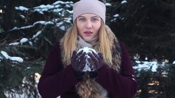 un hermosa niña en un ligero sombrero golpes nieve desde su manos en el abierto aire en el invierno teniendo un bueno estado animico y sonriente video