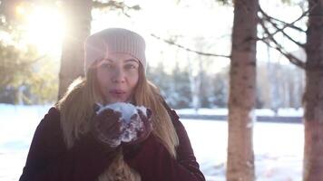 A young cute girl in a light hat blows a pile with snow in the winter in the forest against the background of snow and sun having a good mood. video