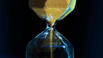 Close up of hourglass with falling golden grains isolated on a dark background. Stock footage. Concept of time, sandglass counting down towards a deadline. video