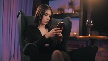 mooi meisje zittend in een fauteuil op zoek Bij de telefoon en drinken wijn in haar huis video