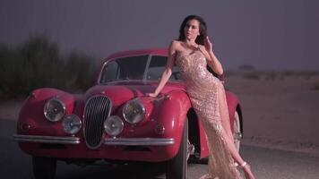 A young woman with long hair and an evening dress stands in high-heeled shoes next to an expensive vintage red car. video