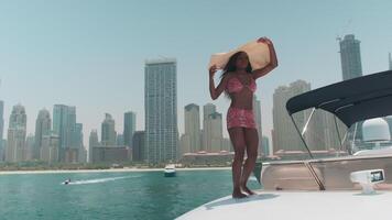 Dubai, VAE - - 5 22 2021. jung dunkel Frau im ein Weiß Bikini und ein Licht Kleid und groß Weiß Hut auf Tafel ein Privat Boot gegen das Hintergrund von das Stadt video