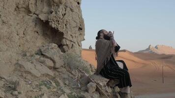 hombre vestido como un chamán, en pie en un rock en el medio de el Desierto playa y jugando el harmónica video