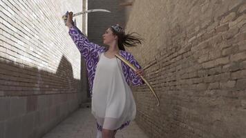 A young woman with long flowing hair, in an oriental robe and skullcap, holds an Asian saber against the backdrop of an ancient city. Slow Motion video