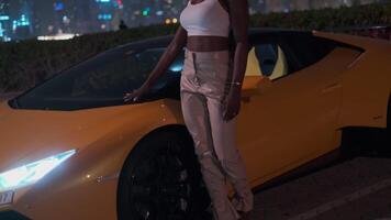 en ung elegant svartmuskig kvinna i högklackade skor står Nästa till ett dyr sporter gul bil på natt video
