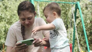 ein jung Mutter mit ein klein Sohn im das Park auf ein sonnig Tag suchen beim das Bildschirm von ein Handy, Mobiltelefon Telefon. video