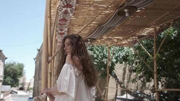 une Jeune femme avec écoulement cheveux dans une lumière blanc robe d'été des stands dans un ancien ville en dessous de une canopée de roseaux video