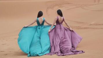 Two young women in long dresses developing in the wind run barefoot through the sand dunes of the desert video