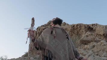 A young bearded man dressed as a shaman with a staff in his hands stands on a lonely rock in the middle of the desert against a clear sky video