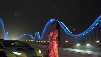 Swarthy young woman in evening dress dancing on a night illuminated bridge next to a sports car video