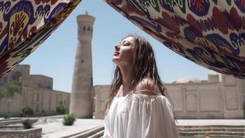 A young woman with flowing hair in a light white sundress stands in the square next to an ancient minaret video