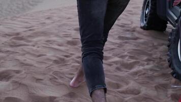 une Jeune homme dans jeans des promenades pieds nus à une petit chariot permanent sur le le sable dans le désert video
