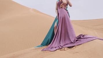 Two young women in long dresses developing in the wind stand barefoot through the sand dunes of the desert video