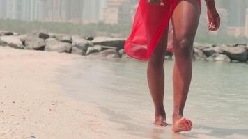 A young swarthy woman in a bikini and a light red dress walks barefoot on the rocks against the backdrop of the sea and skyscrapers of Dubai. video