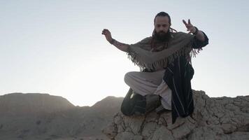 uma jovem jovem xamã dentro a lótus posição ora em uma Rocha contra a pano de fundo do pôr do sol entre a areia dunas do a deserto video