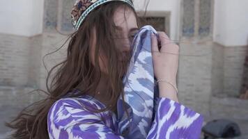 A young woman with long hair in an oriental robe and skullcap stands in the wind near an ancient mosque video