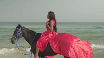 un joven moreno mujer en un rojo vestir se sienta en un caballo en pie en el Oceano costa. video