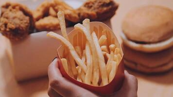 Closeup image of a woman holding and eating french fries and hamburger with fried chicken on the table at home video