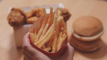 avvicinamento Immagine di un' donna Tenere e mangiare francese patatine fritte e Hamburger con fritte pollo su il tavolo a casa video