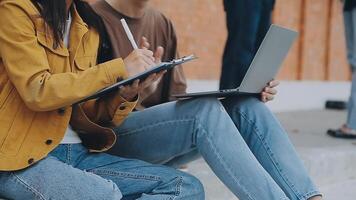 Group of happy young Asian college students sitting on a bench, looking at a laptop screen, discussing and brainstorming on their school project together. video