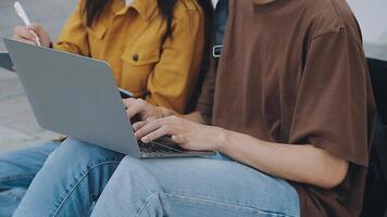 Group of happy young Asian college students sitting on a bench, looking at a laptop screen, discussing and brainstorming on their school project together. video