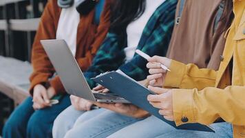 Group of happy young Asian college students sitting on a bench, looking at a laptop screen, discussing and brainstorming on their school project together. video