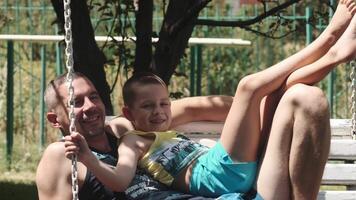 A teenager lies on top of his young father on a swing in a park on a sunny day. They talk and laugh video