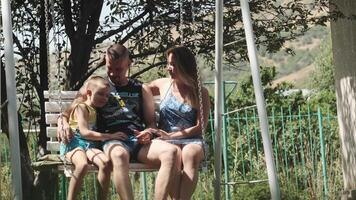 familia. padre, madre y hijo balanceo en un columpio en el parque en un soleado día. ellos abrazo y reír. video