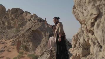 ein jung bärtig Mann gekleidet wie ein Schamane mit ein Mitarbeiter im seine Hände steht auf ein einsam Felsen im das Mitte von das Wüste gegen ein klar Himmel video