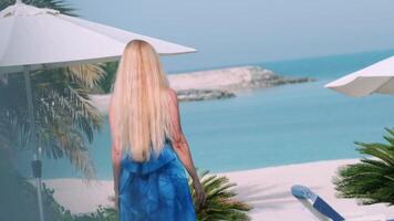 blond Mädchen tragen lange Blau Sommer- Kleid Spaziergänge auf das Terrasse mit tropisch Pflanzen video