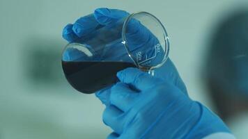 un' laboratorio tecnico nel un' bianca cappotto e protettivo guanti versa liquido in un' chimico piatto. video