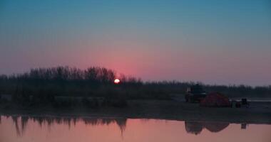 The SUV stands next to a stretched awning on the shore of the lake, surrounded by trees at sunset video