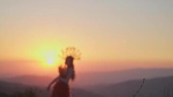 giovane femmina modello nel progettista copricapo fatto di spighette e vestito fatto di fiori e erba su il sfondo di tramonto nel il montagne. video