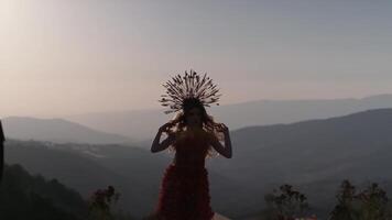 A young woman in an autumn outfit of flowers and grass and a headdress made of ears of corn stands against the background of the sunset in the mountains video