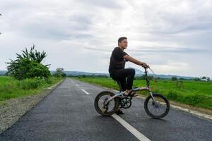 contento asiático hombre montando un bicicleta a el Mañana en el asfalto la carretera. ciclismo con montaña y arrozal arroz campo ver a el antecedentes. foto