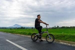 contento asiático hombre montando un bicicleta a el Mañana en el asfalto la carretera. ciclismo con montaña y arrozal arroz campo ver a el antecedentes. foto