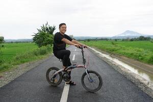 contento asiático hombre montando un bicicleta a el Mañana en el asfalto la carretera. ciclismo con montaña y arrozal arroz campo ver a el antecedentes. foto