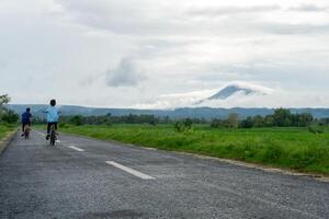 Two Asian kids riding bicycle at the morning on the asphalt road. Two boys cycling with mountain and paddy rice field view at the background. copy space photo