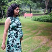 un embarazada indio dama poses para al aire libre el embarazo disparar y manos en barriga, indio embarazada mujer pone su mano en su estómago con un maternidad vestir a sociedad parque, embarazada fuera de maternidad disparar foto