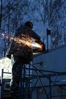 A construction worker works on a construction site, stands on scaffolding, holds an electric cutting machine, sparks fly, a trail of sparks, dark, low angle view photo