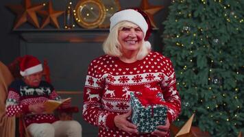 Senior grandmother in festive sweater presenting Christmas gift box, smiling, looking at camera video