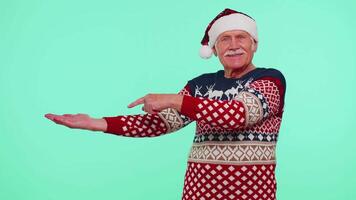 Grandfather in Santa Christmas sweater showing thumbs up and pointing empty place, advertising area video