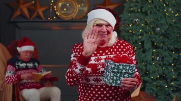 Senior grandmother in Christmas sweater smiling friendly at camera and waving hands gesturing hello video