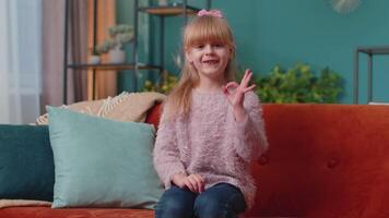 retrato do pequeno criança menina criança sozinho em sofá às casa mostrando Está bem gesto, gostar sinal, positivo Boa video