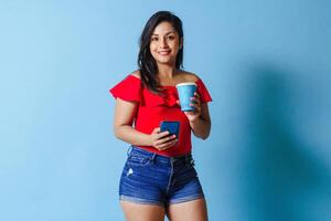 retrato joven latinx mujer utilizando móvil teléfono participación para llevar café taza Mira a cámara. foto