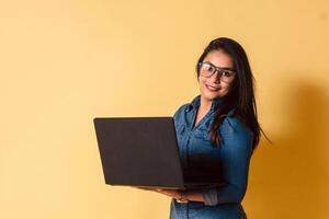 Portrait of young woman with glasses holding laptop smiling look camera over yellow background. photo