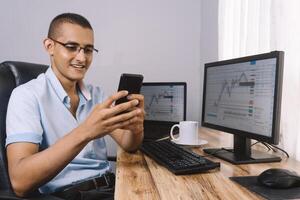 young Hispanic man using his mobile phone in front of his computer. portrait of young investor and cheerful trader watching his smartphone. photo