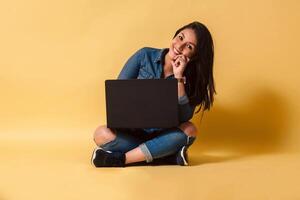 Full length of a woman holding laptop looking at camera sitting on floor over yellow background. photo